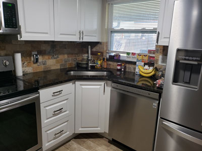 The "After" remodeling picture of a kitchen. White cabinets, black granite countertops, stainless steel appliances, tile backsplash, porcelain wood-look tile flooring