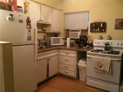 The "Before" remodeling picture of a kitchen: Old white appliances, formica countertops, sink wedged against fridge, wooden floor, no backsplash.